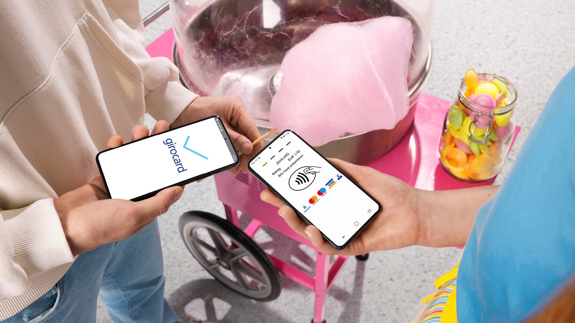 Zwei Personen führen eine kontaktlose Zahlung per Smartphone durch, eines zeigt das Girocard-Logo, das andere eine Zahlungsbestätigung. Im Hintergrund steht eine pinke Zuckerwattenmaschine und ein Glas mit bunten Lutschern.